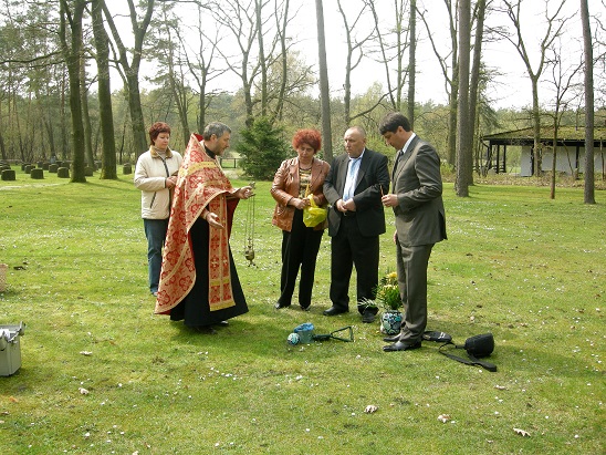 Fünf Menschen stehen auf einer Wiese im Kreis um ein Objekt auf dem Boden. Einer trägt Priesterkleidung, er scheint etwas zu segnen.