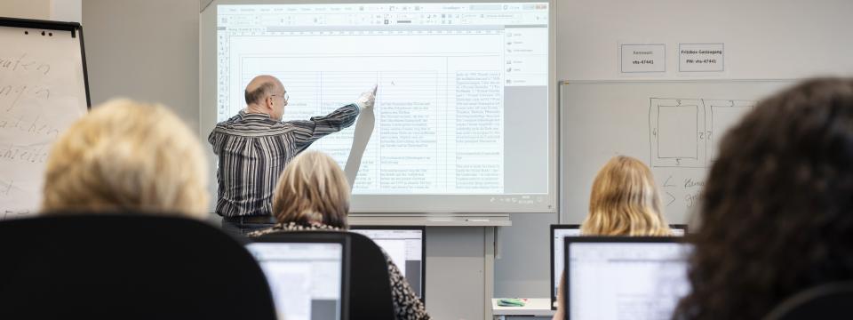 Mehrere Personen sitzen an Computern. Sie hören einem Mann zu, der vorne eine Präsentation erläutert.