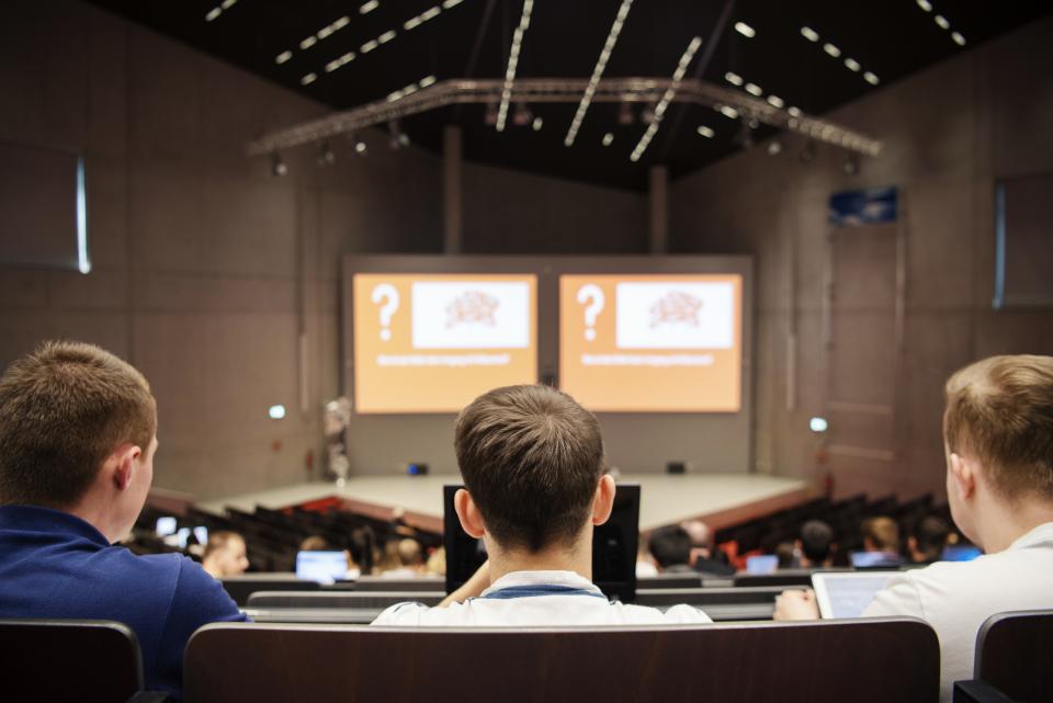 In einem Hörsaal schauen drei junge Menschen nach vorne auf den Vortrag des Dozierenden. Auf der Präsentation vorne sind Fragezeichen eingeblendet.