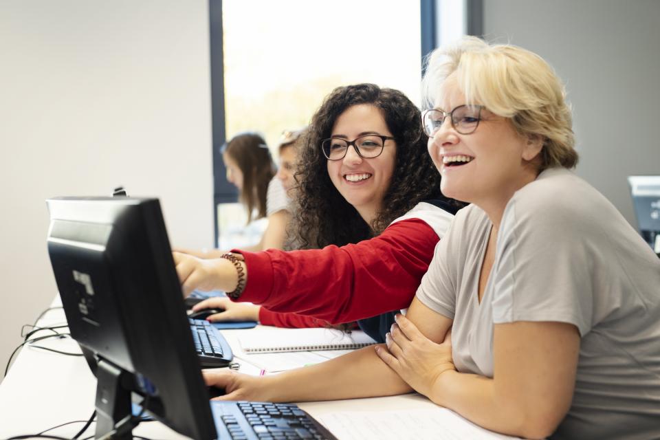 Eine jüngere und eine ältere Frau am Computer, die jüngere Frau zeigt der anderen etwas.