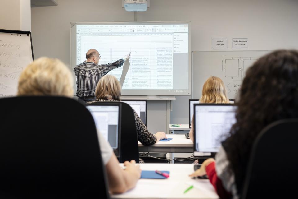 Mehrere Personen sitzen an Computern. Sie hören einem Mann zu, der vorne eine Präsentation erläutert.